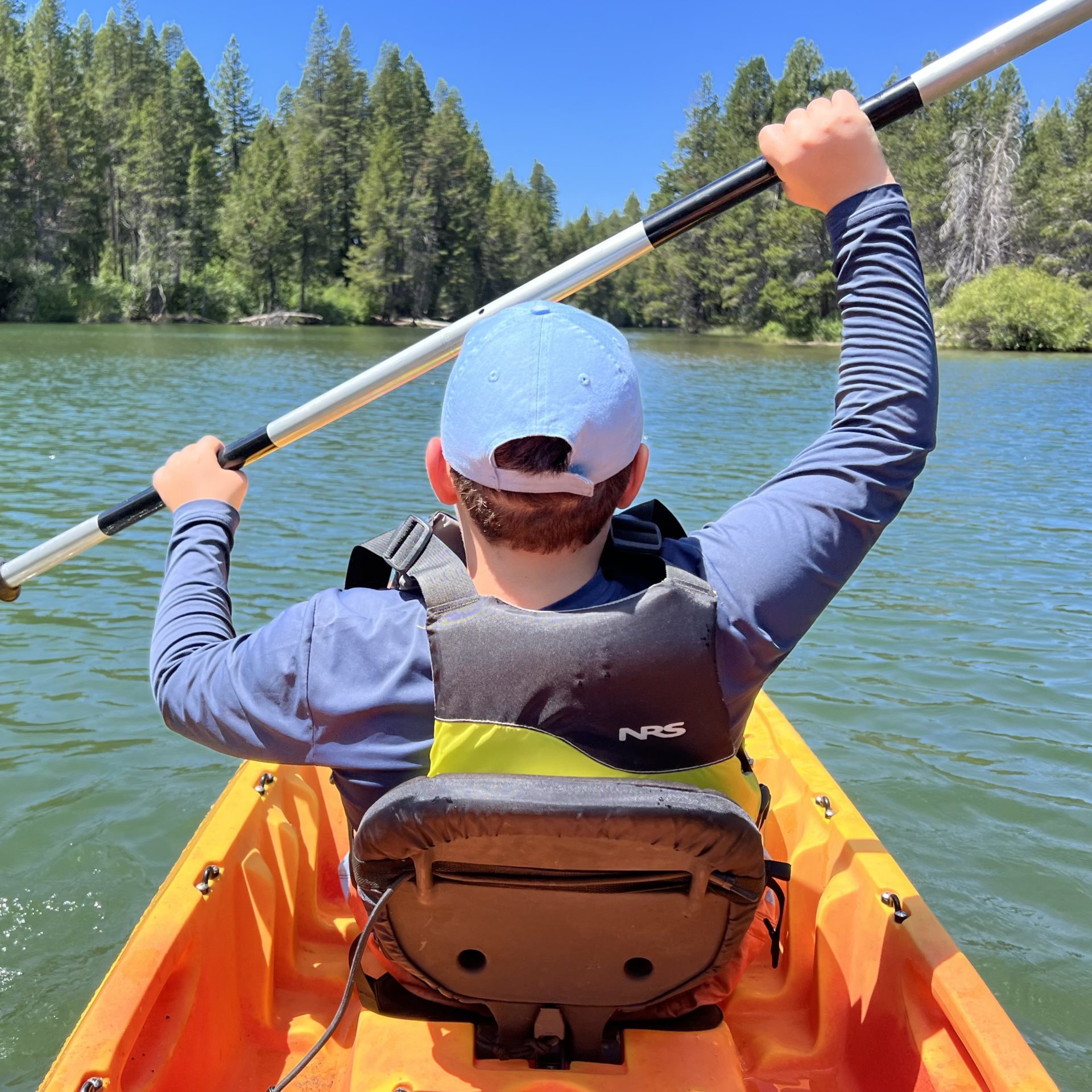 Boy in blue baseball cap and shirt paddling orange kayak in alpine lake